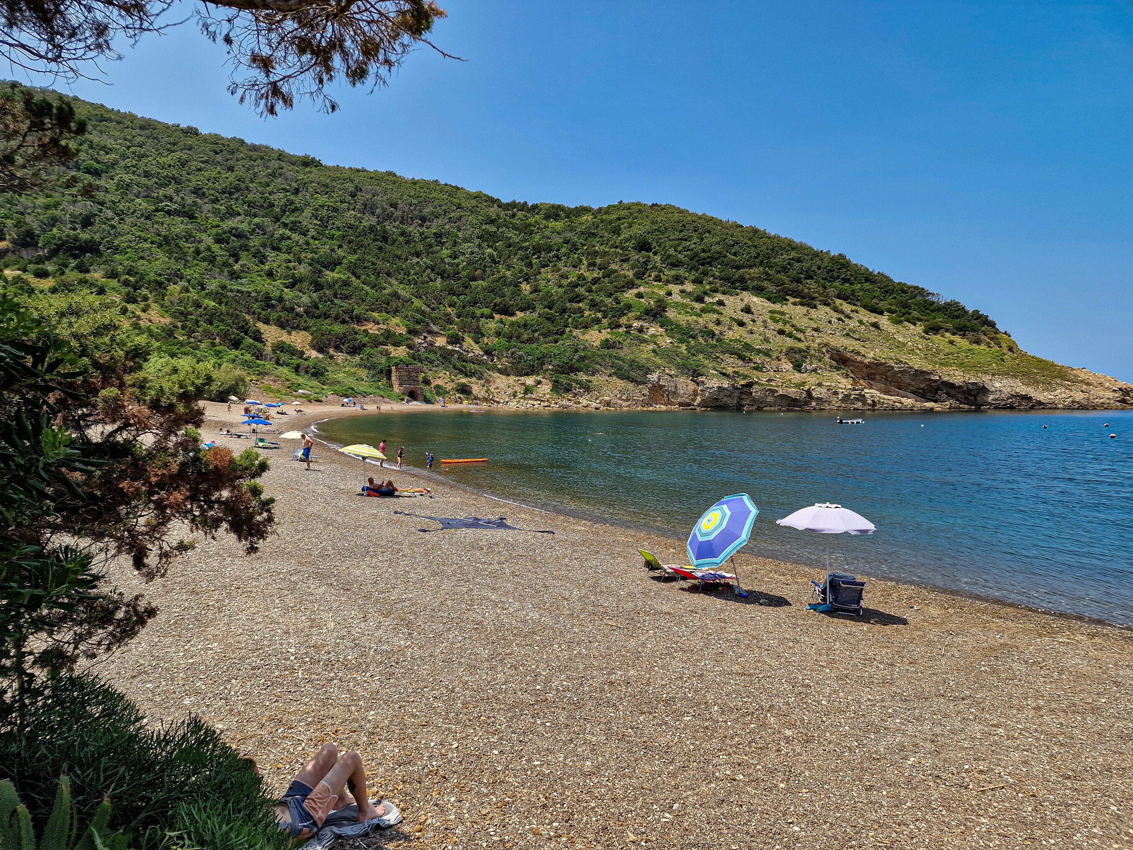 Une plage de galets avec des parasols, un ciel dégagé et une colline en arrière-plan.