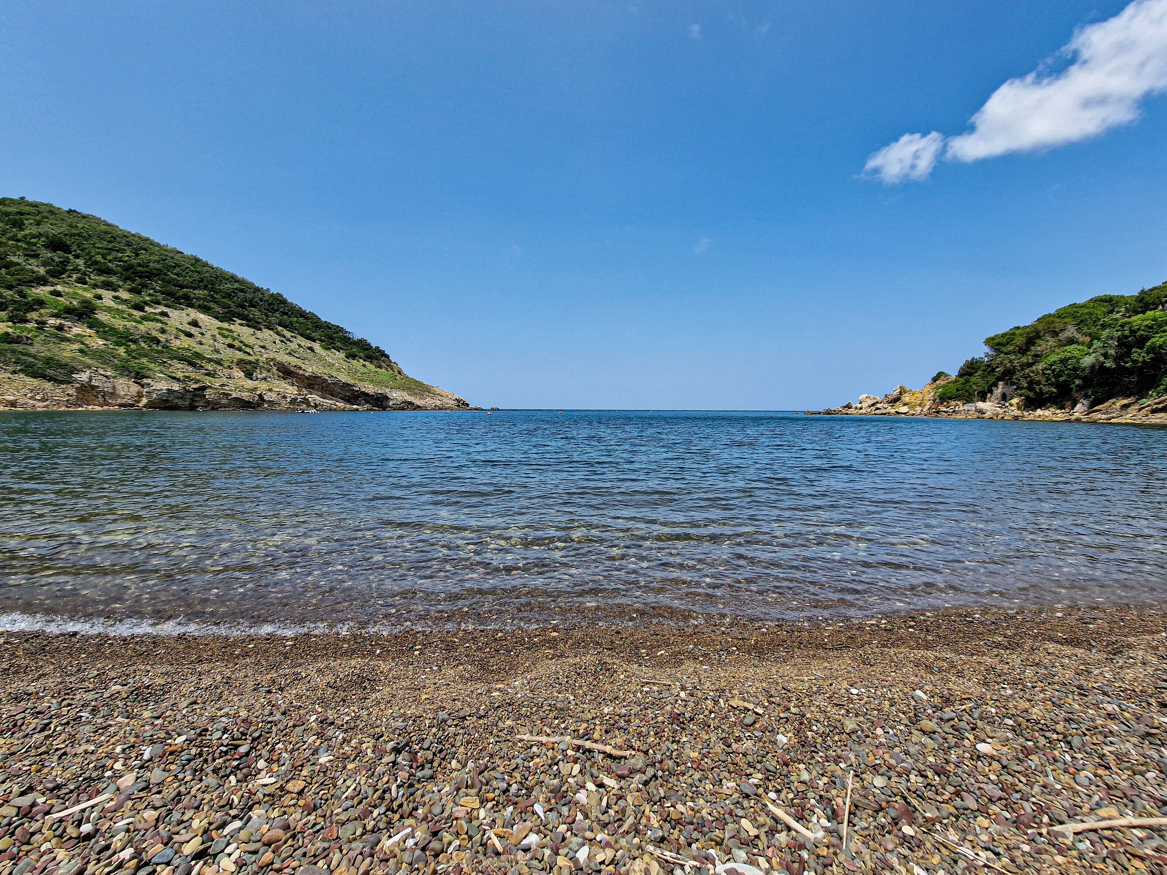 A pebbled beach with clear water and hills in the background.