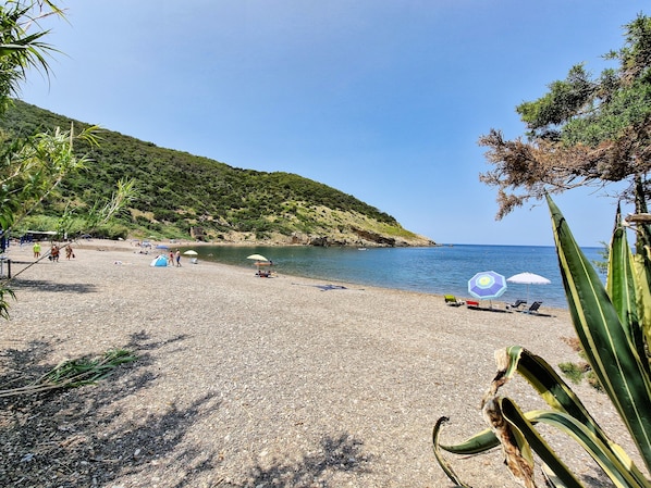 Una playa de guijarros con sombrillas, cielo despejado y un paisaje montañoso al fondo.