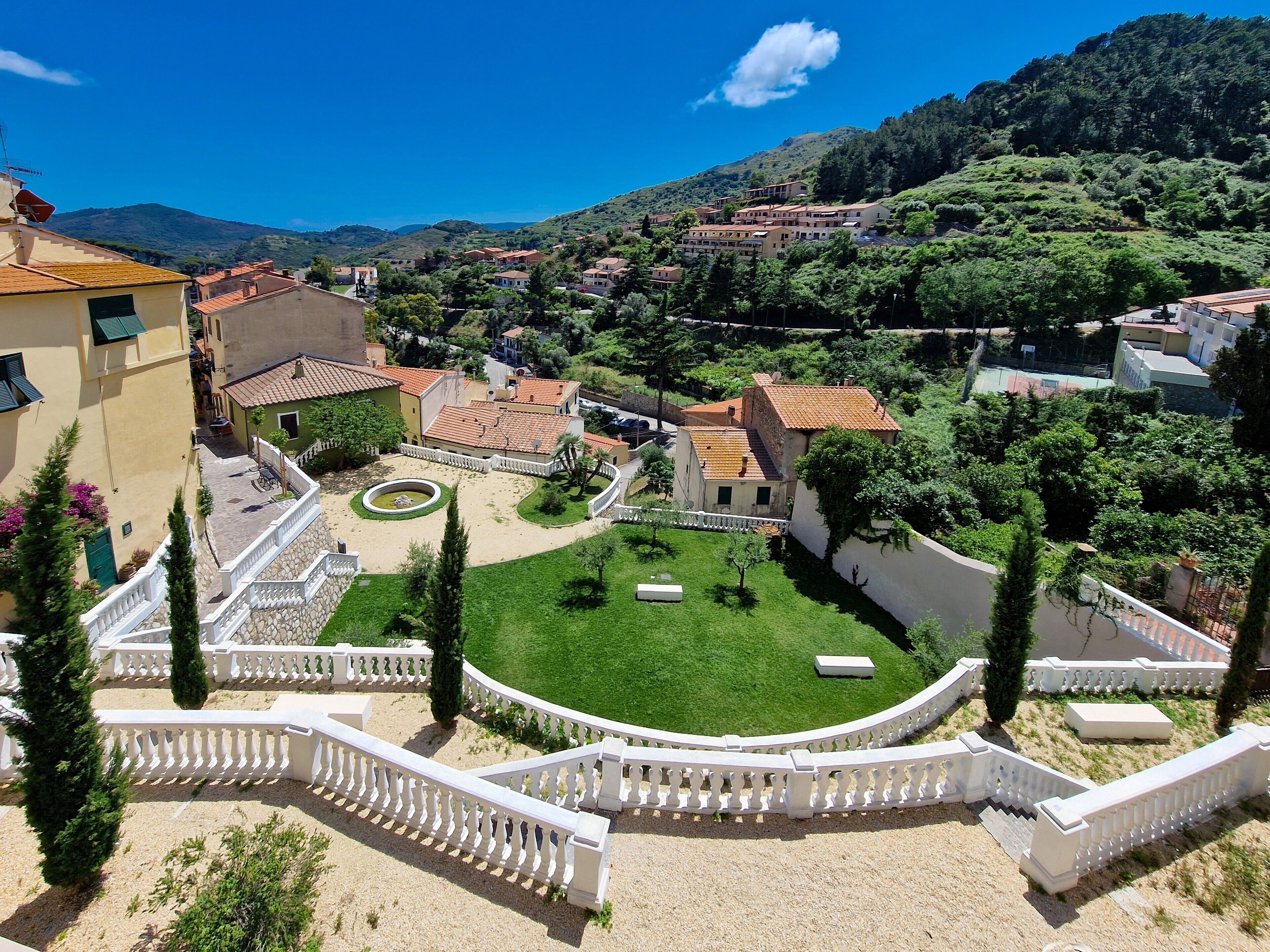 A courtyard with a fountain, surrounded by buildings with red-tiled roofs, and a mountainous backdrop.