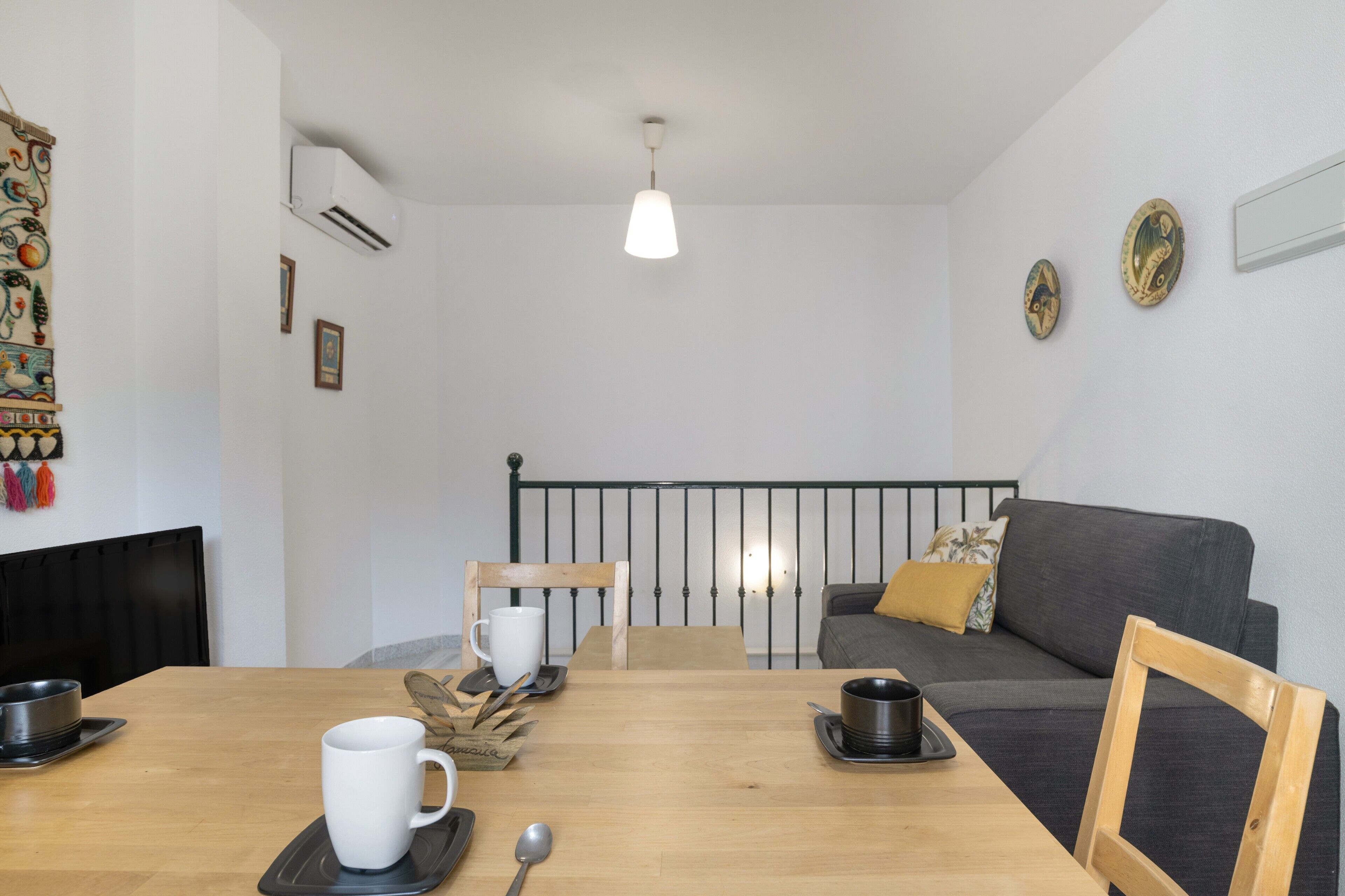 A room with a wooden dining table, a gray sofa, a black metal bed frame, and a hanging light fixture.