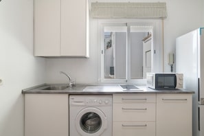 A compact kitchen with a washing machine, a microwave, a sink, and a window with a view of the street.