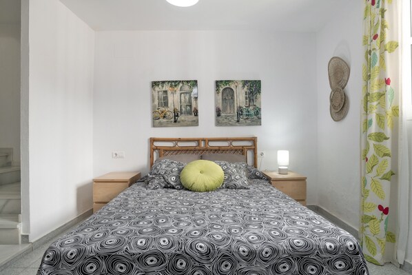 A bedroom with a patterned bedspread, a wooden headboard, two wall-mounted paintings, a nightstand, a lamp, and a window with a floral curtain.