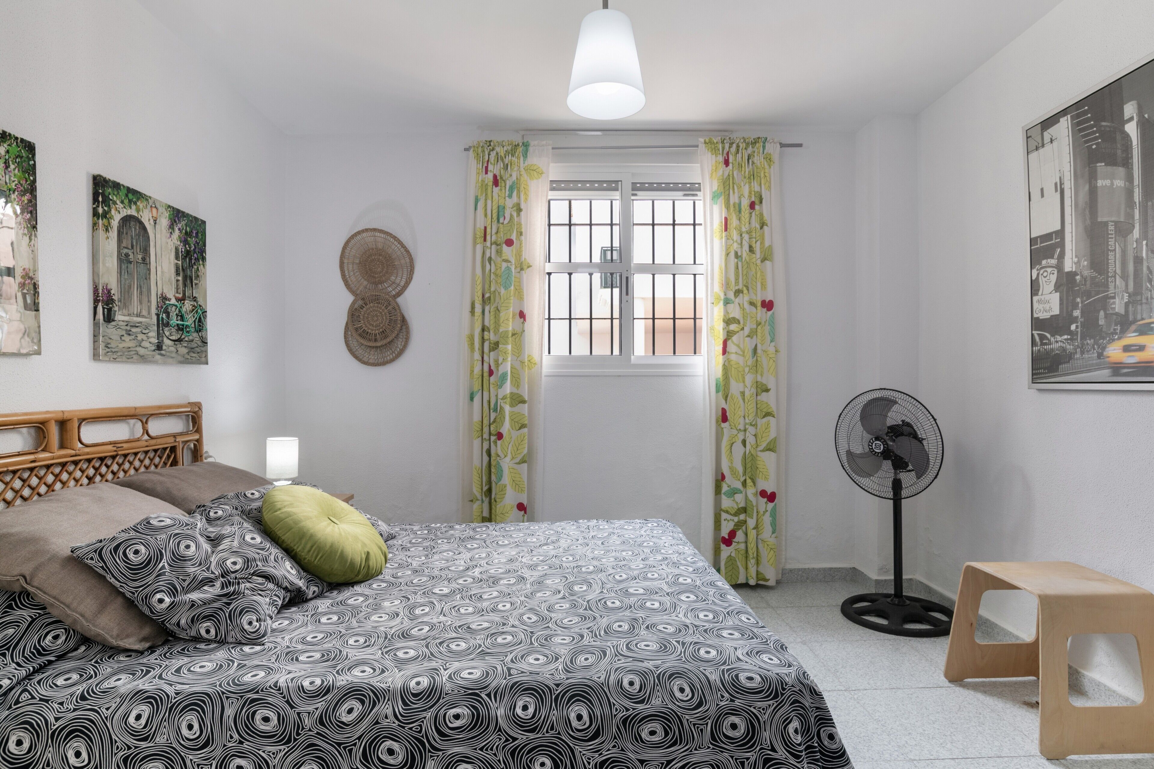 Une chambre à coucher avec un lit, une tête de lit en bois, un ventilateur sur pied, une fenêtre avec des rideaux et des œuvres d’art murales.