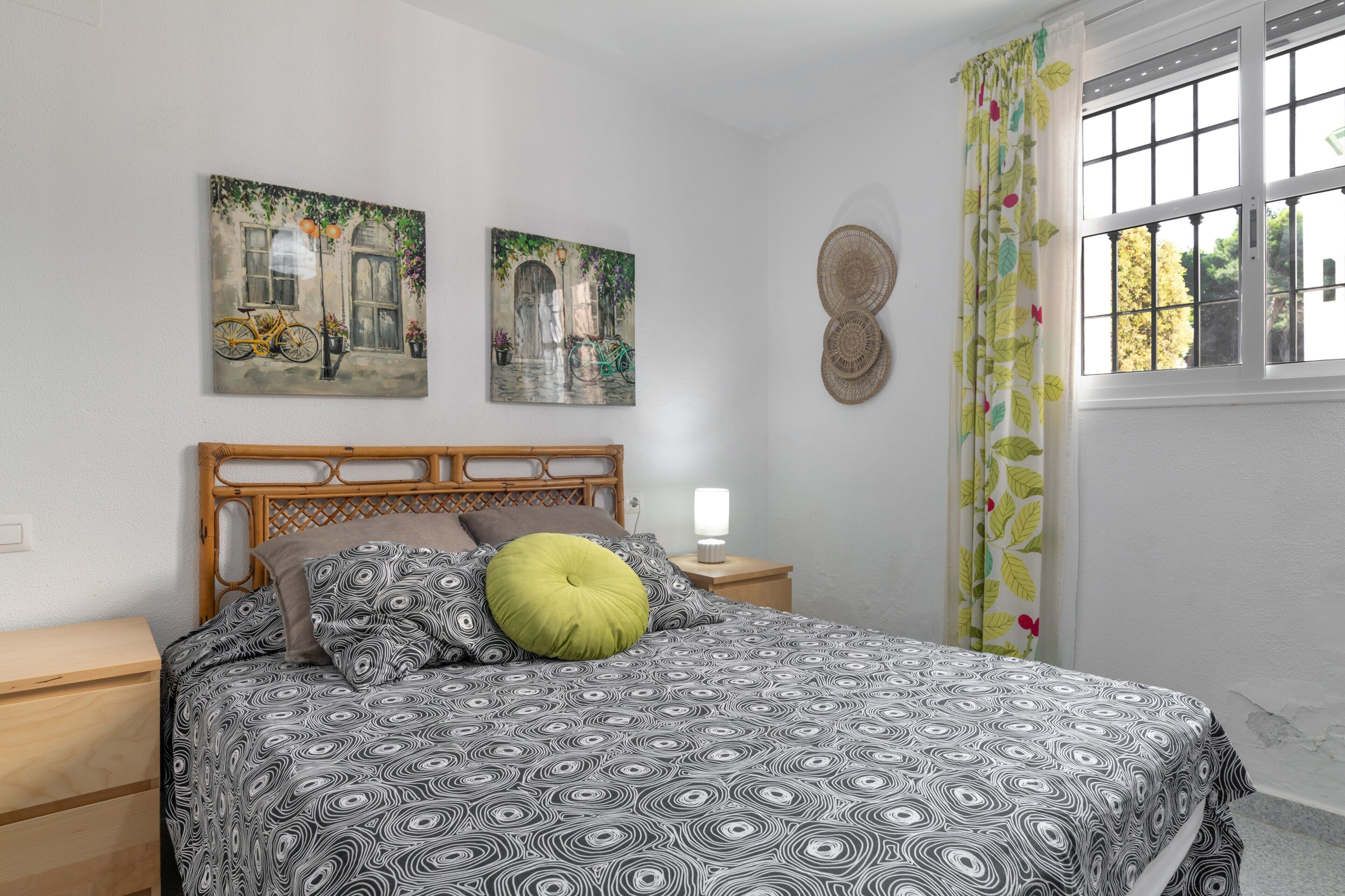 A bedroom with a patterned bedspread, a wooden headboard, a nightstand with a lamp, and two wall-mounted paintings.