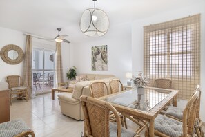 Un vaste espace de vie comprenant une table à manger avec un plateau en verre, des meubles en osier, un ventilateur de plafond et une grande fenêtre offrant une vue dégagée.