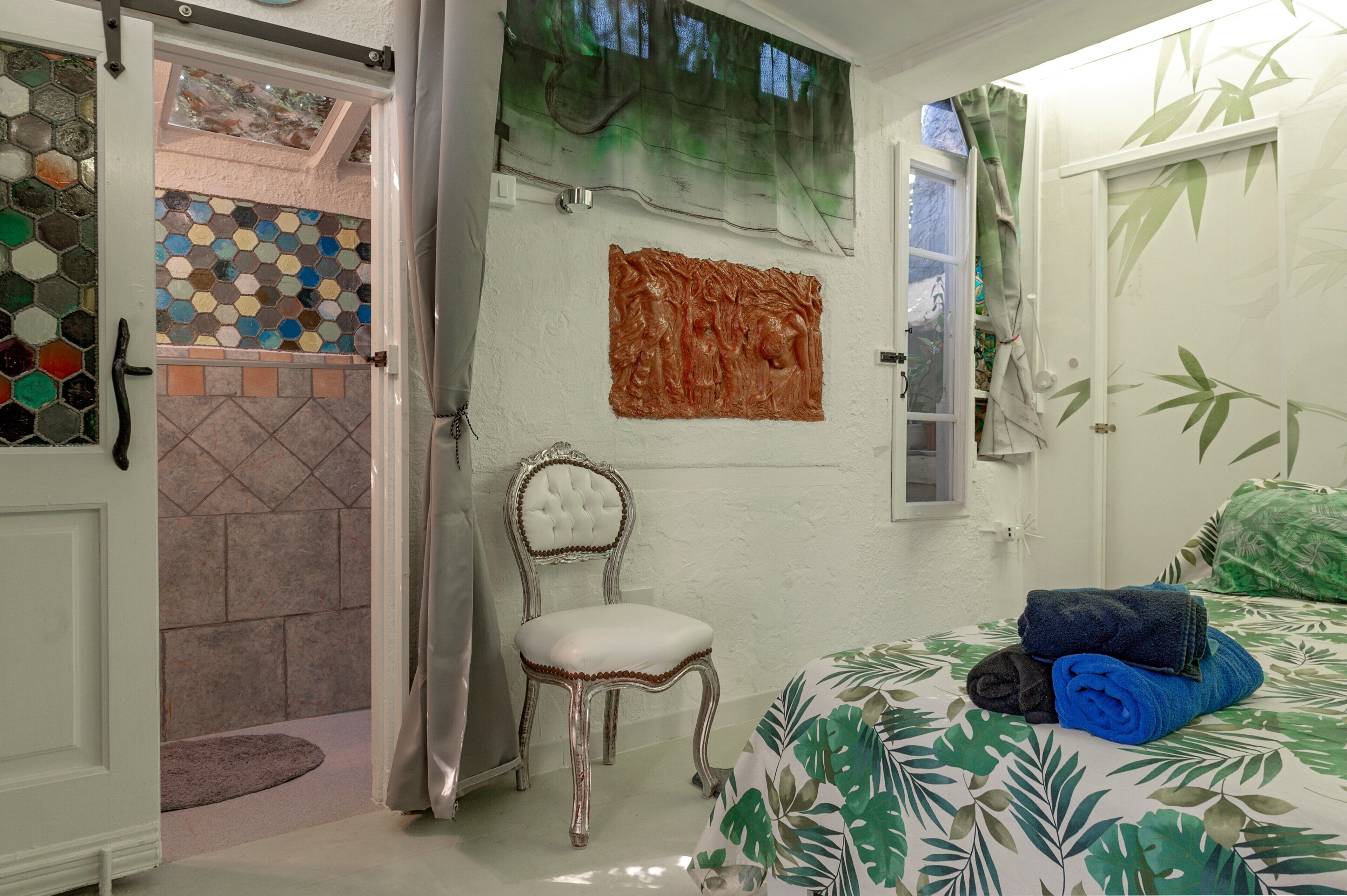 A room with a bed, a chair, a door, and a wall with a mosaic and a painting.