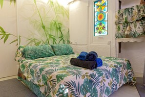 A bed with a tropical leaf pattern bedspread, a pair of folded towels, a bamboo wall mural, and a colorful stained glass window.