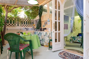 Um quarto com uma cama, uma mesa com cadeiras, uma janela com cortinas e uma porta que dá para outro cômodo.