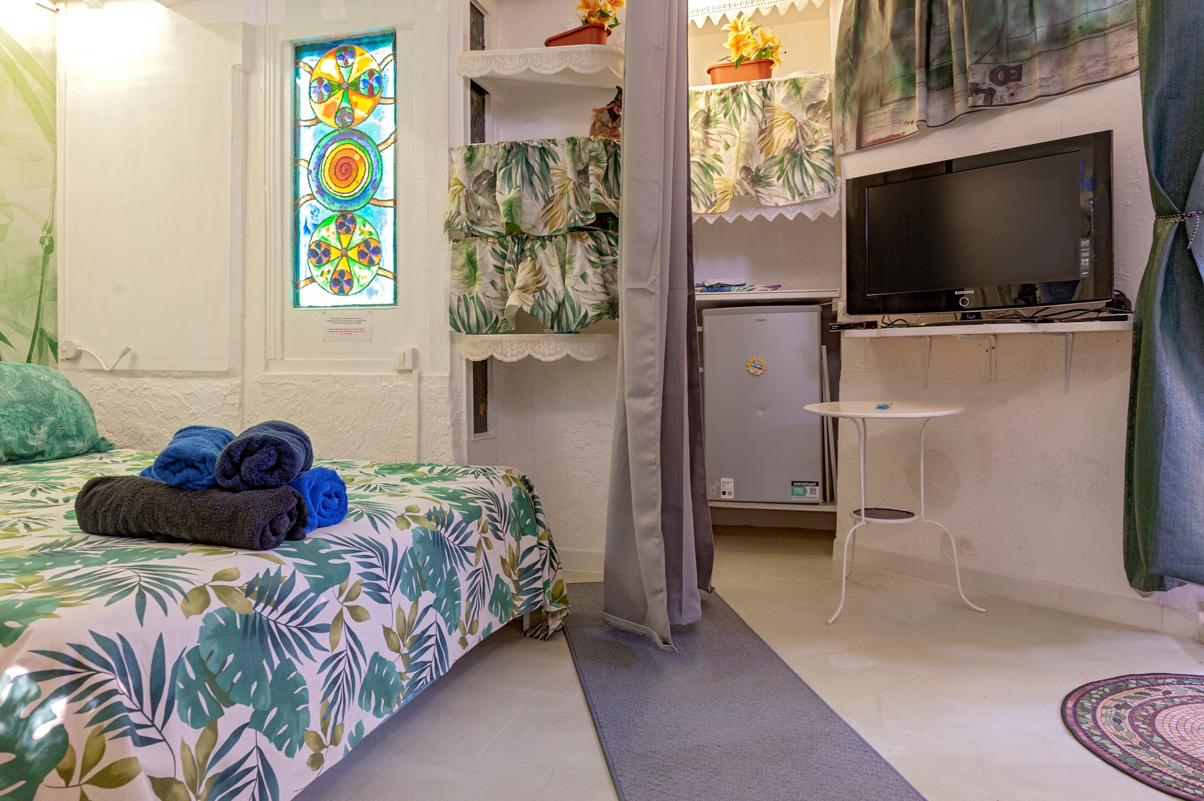 Una habitación con cama, televisor, refrigerador pequeño y ventana con diseño de vitral.