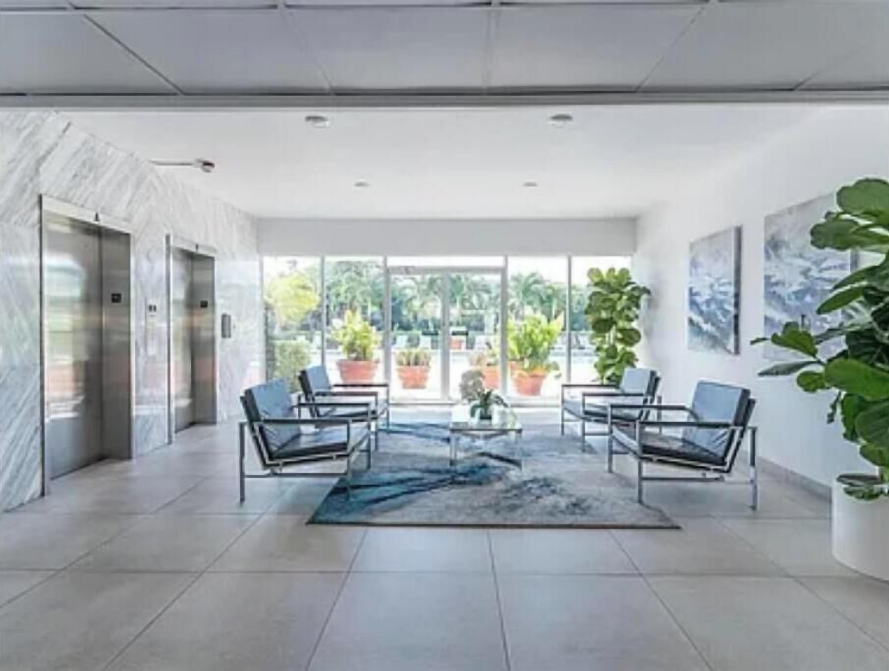 Un hall d’entrée moderne avec un sol en marbre, deux ascenseurs, un espace détente avec des fauteuils, une table basse et de grandes fenêtres donnant sur l’extérieur.