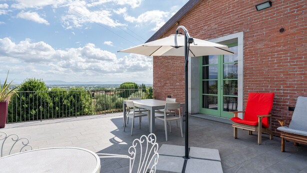 A patio with a table and chairs, a red chair, a white umbrella, and a brick building with a green door. - Holiday Home 'Dulù - Luminoso Spazio' with Shared Garden, Balcony and Wi-Fi (Capannori)