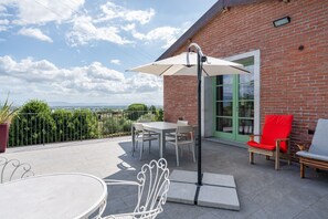 Une terrasse avec une table et des chaises, une chaise rouge, un parasol blanc et un bâtiment en briques avec une porte verte.