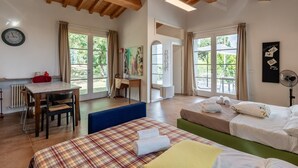 A room with wooden beams, a bed with a plaid bedspread, a desk with a chair, and a view of greenery outside. - Holiday Home 'Dulù - Luminoso Spazio' with Shared Garden, Balcony and Wi-Fi (Capannori)