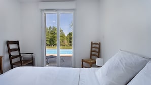 A bedroom with a bed, two wooden chairs, a white lamp, and a sliding glass door leading to a pool area.
