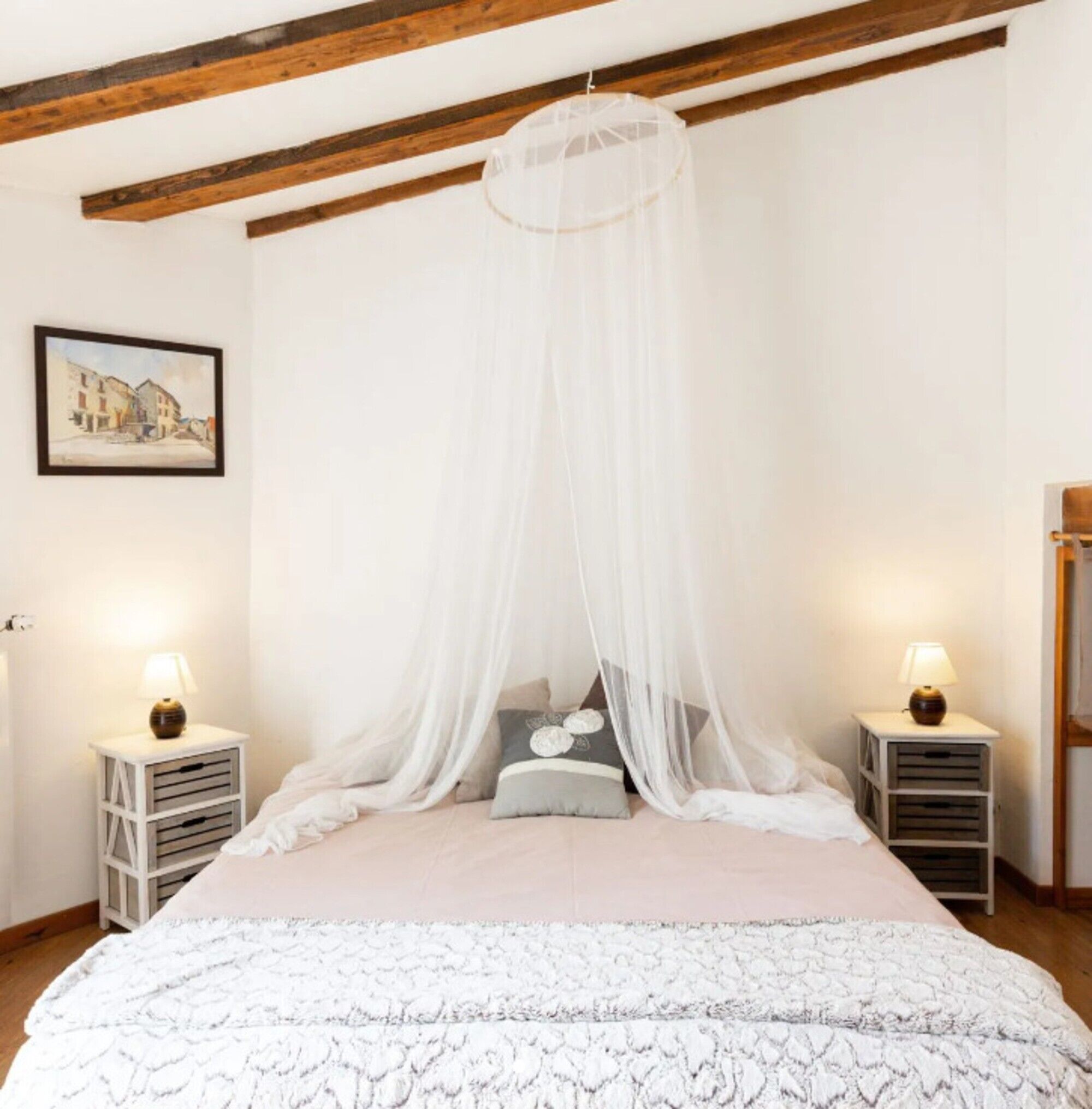 A bedroom with a canopy bed, bedside tables with lamps, a framed picture on the wall, and a wooden ceiling.