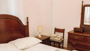Une chambre à coucher avec un lit en bois, une table de chevet avec une lampe, une chaise sur laquelle sont pliés des vêtements et une commode avec un miroir.