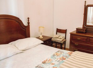 Quarto com cama de madeira, mesa de cabeceira com abajur, cadeira com roupas dobradas e cômoda com espelho.