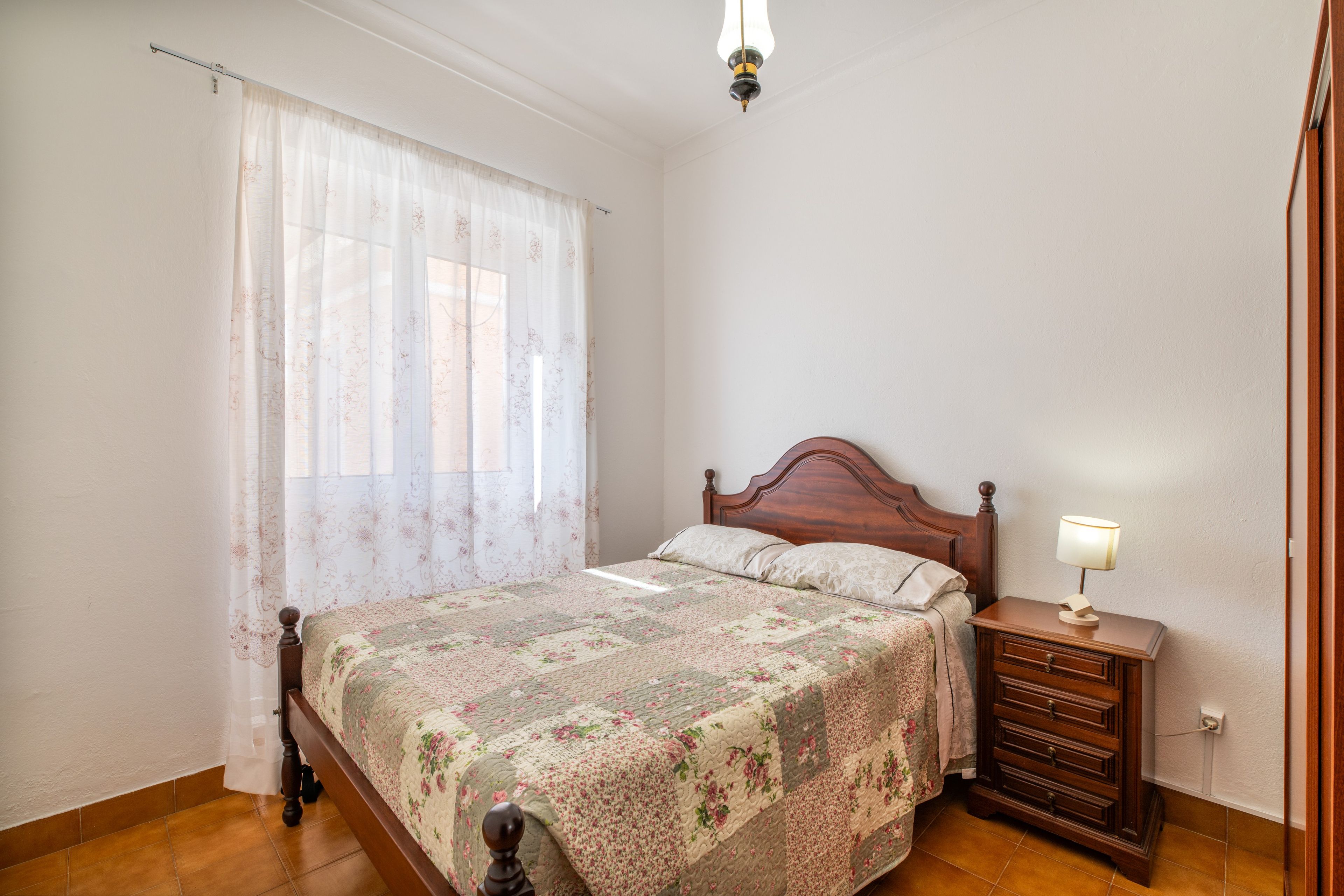 A bedroom with a wooden bed, a nightstand with a lamp, and a window with sheer curtains.