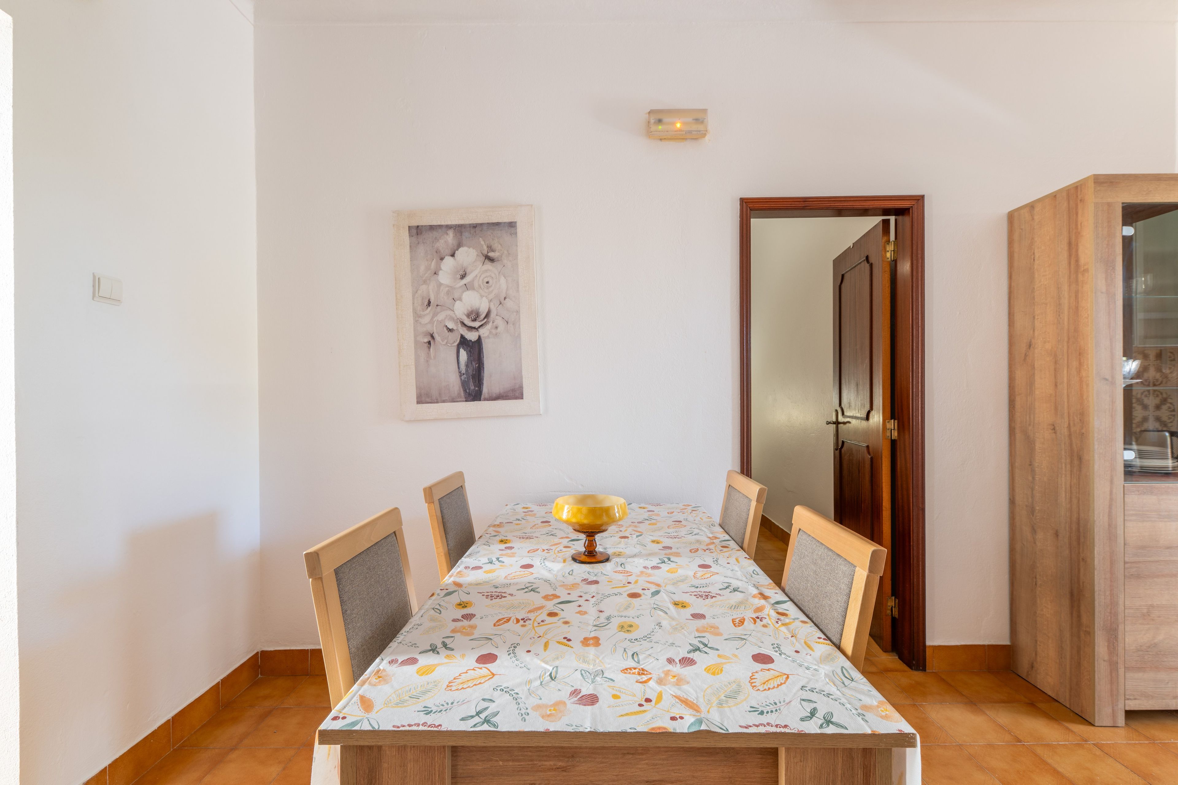 A dining area with a table set for four, a vase of flowers on the wall, and a wooden cabinet to the right.