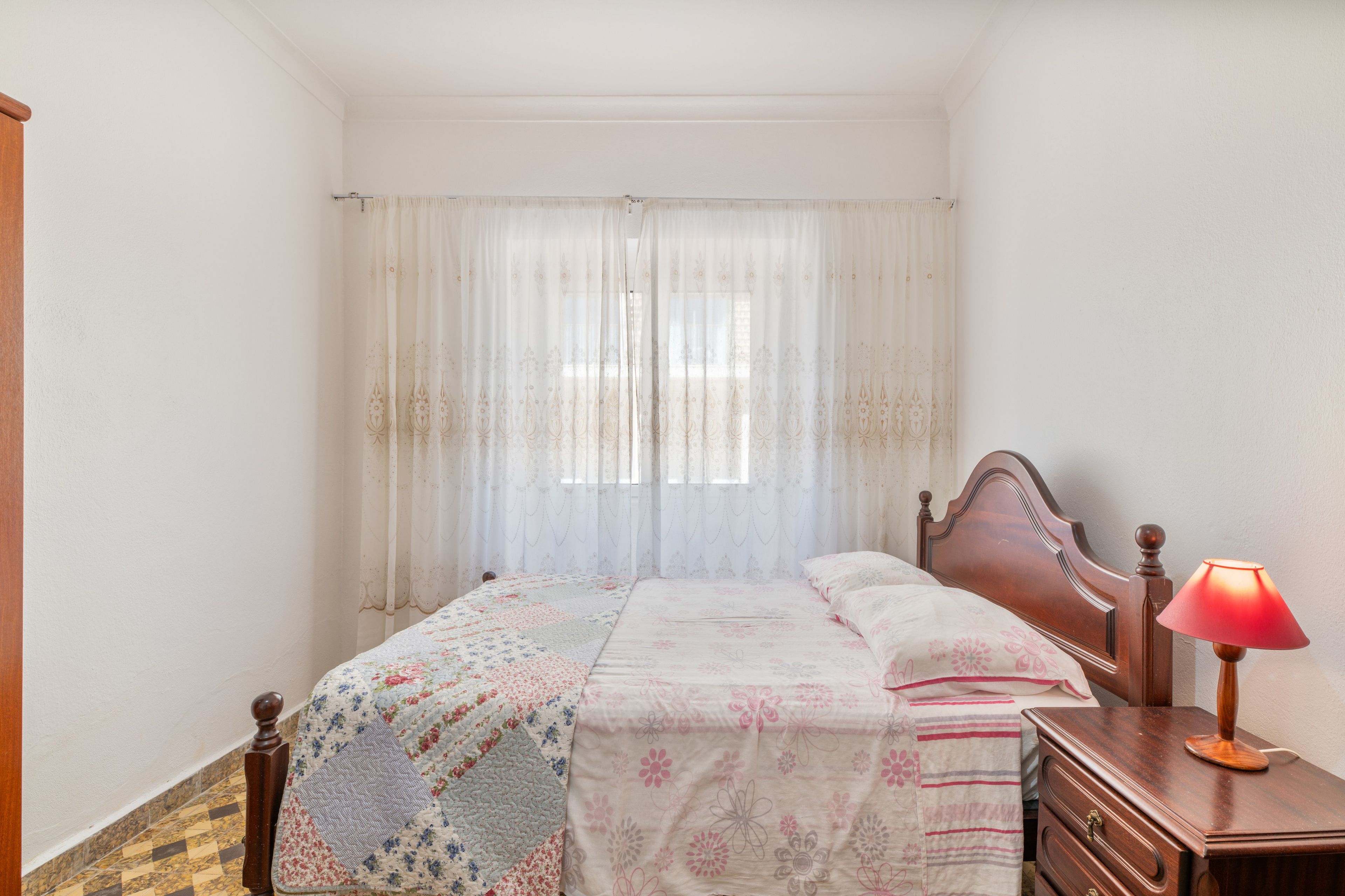 A bedroom with a bed, a wooden dresser, a lamp, and a window with sheer curtains.