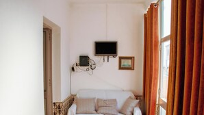 Une pièce avec un canapé blanc, un téléviseur à écran plat fixé au mur, un tableau encadré et une fenêtre avec des rideaux.