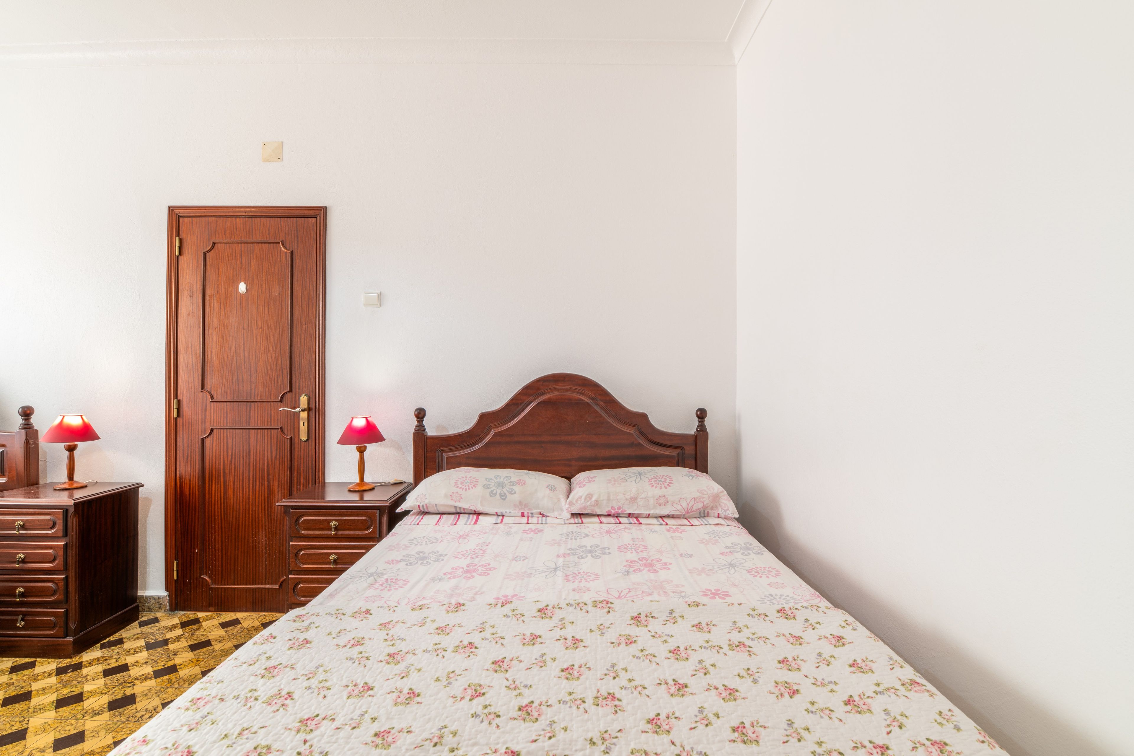 Ein Schlafzimmer mit Holztür, einem Bett mit Blumenmuster-Bettwäsche, einem Nachttisch mit Lampe und einer Kommode mit Schubladen.