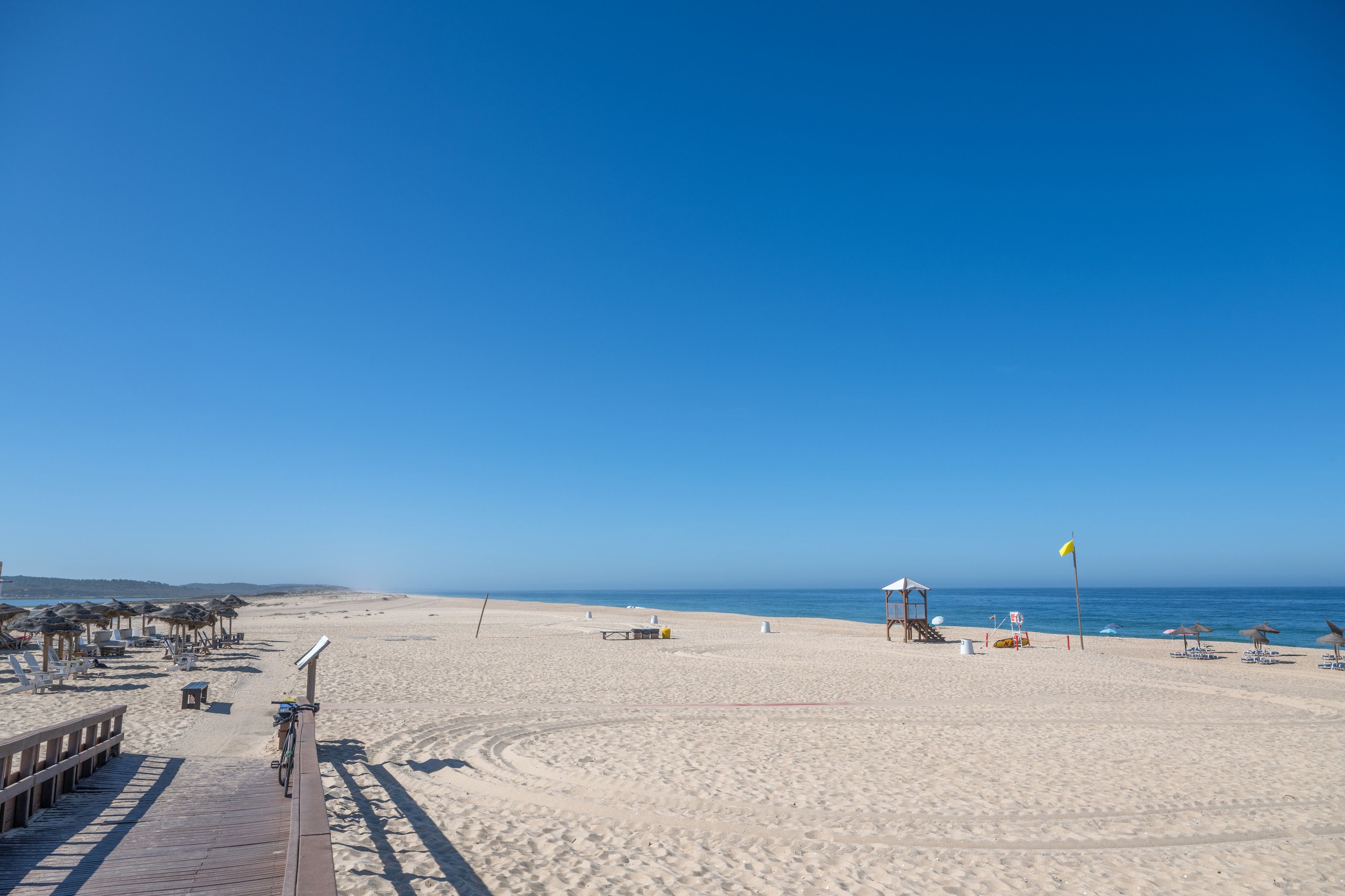 Ein Strand mit einem hölzernen Promenadenweg, Liegestühlen, Sonnenschirmen und einem Rettungsschwimmerstand.