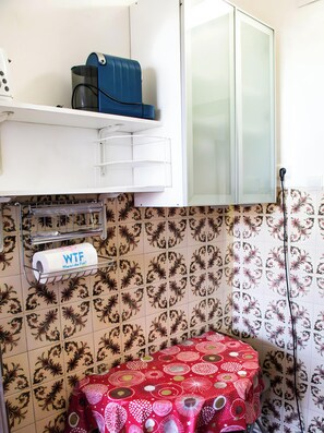 Een badkamer met tegelvloer, een toilet met een rode, bewerkte zitting, een handdoekhouder en een rol toiletpapier, en een kastje met een blauw voorwerp erboven.