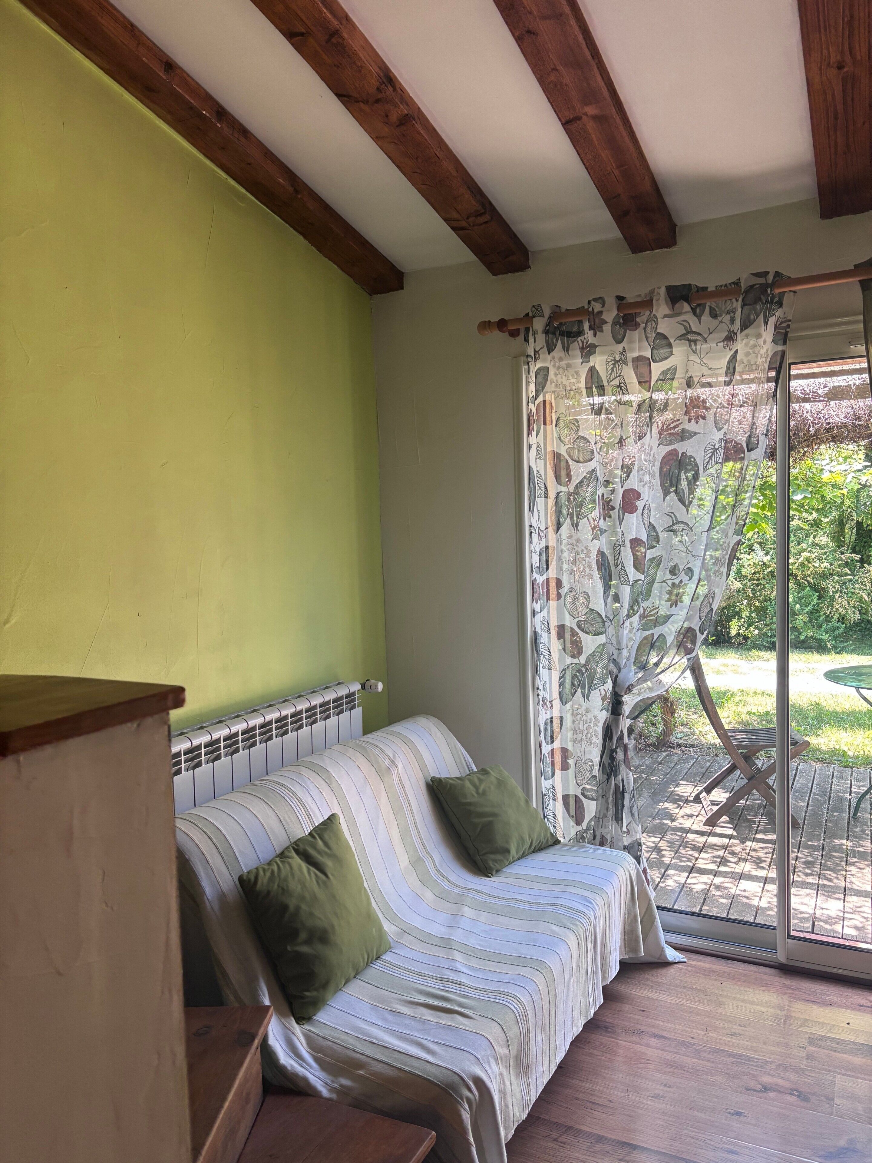 A room with a green wall, a striped sofa with green cushions, a wooden table, and a glass door leading to an outdoor area with a table and chairs.