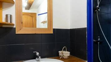Un baño con tocador de madera, un lavabo blanco, un espejo, cabina de ducha y una pequeña cesta de mimbre.