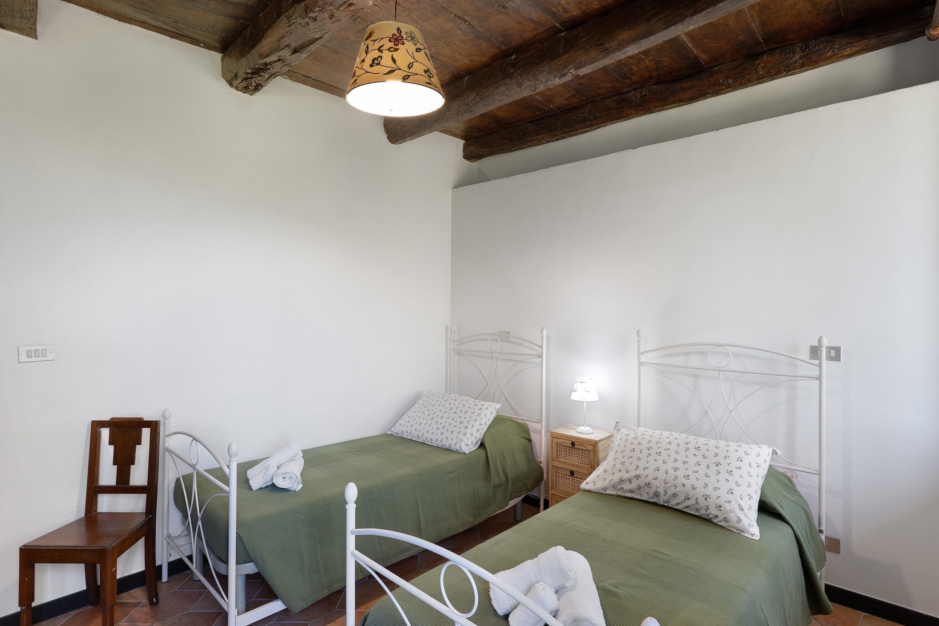 A bedroom with two beds, a wooden chair, a bedside table with a lamp, and a hanging light fixture.