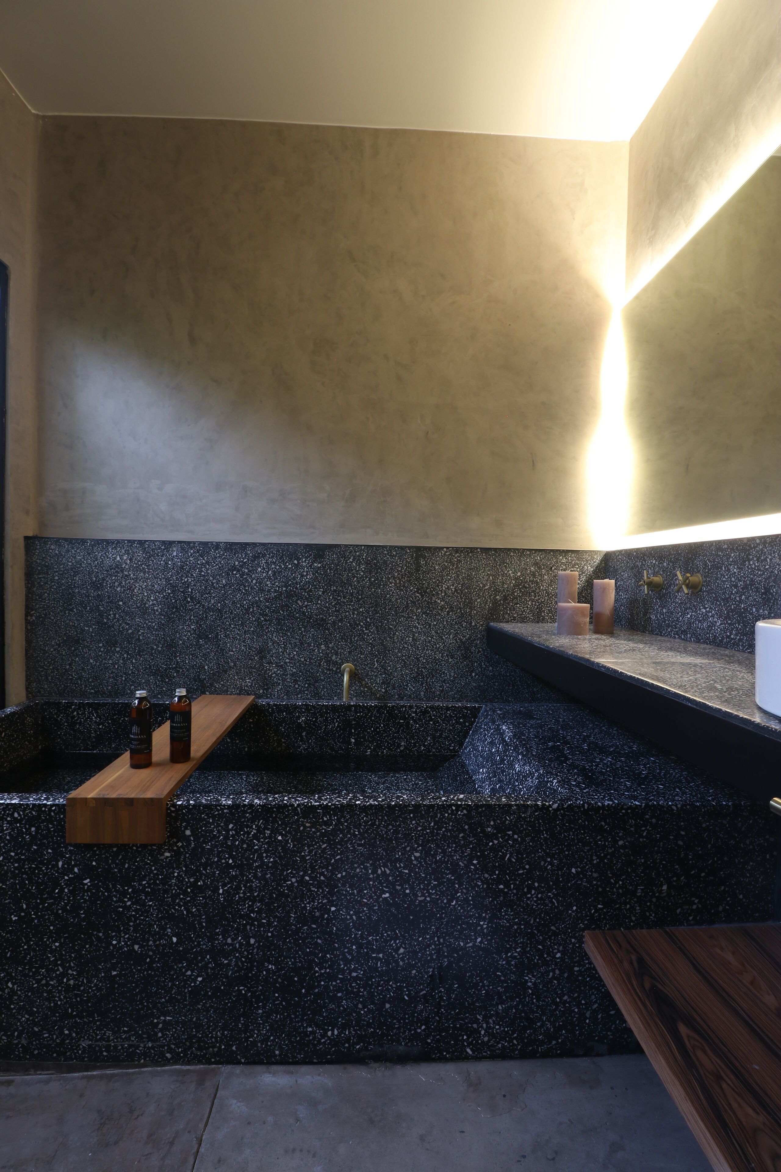 A modern bathroom with a black speckled countertop, wooden shelves, and a wall-mounted light fixture.