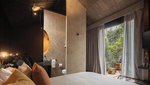 A modern hotel room with a large bed, a round mirror, a bathroom with a toilet, and a view of the outdoors through large windows.