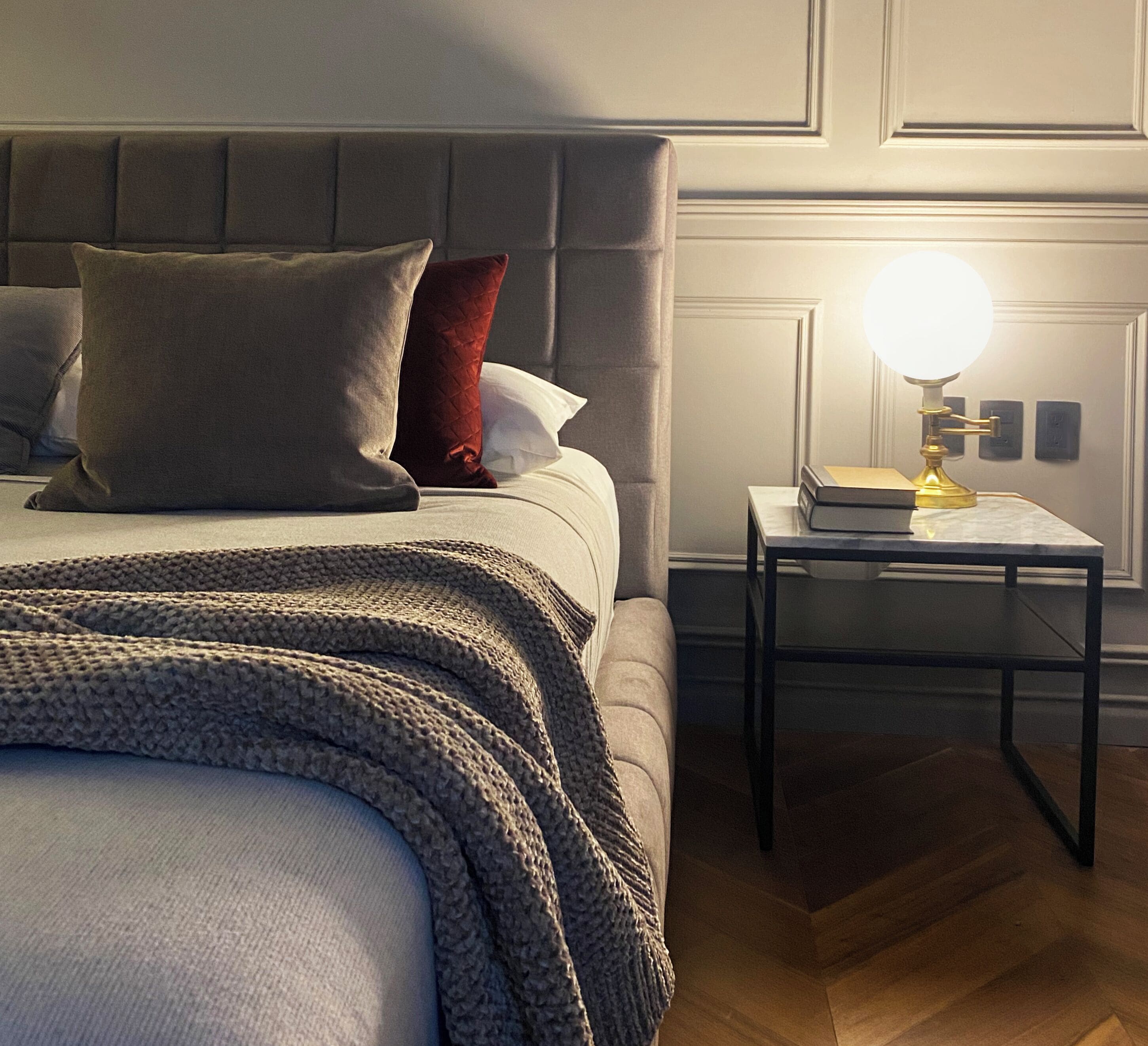 Un lit bien fait, avec une tête de lit grise, un oreiller rouge et une couverture grise. Une petite table de chevet, sur laquelle se trouvent une lampe et un livre, est placée à côté du lit.