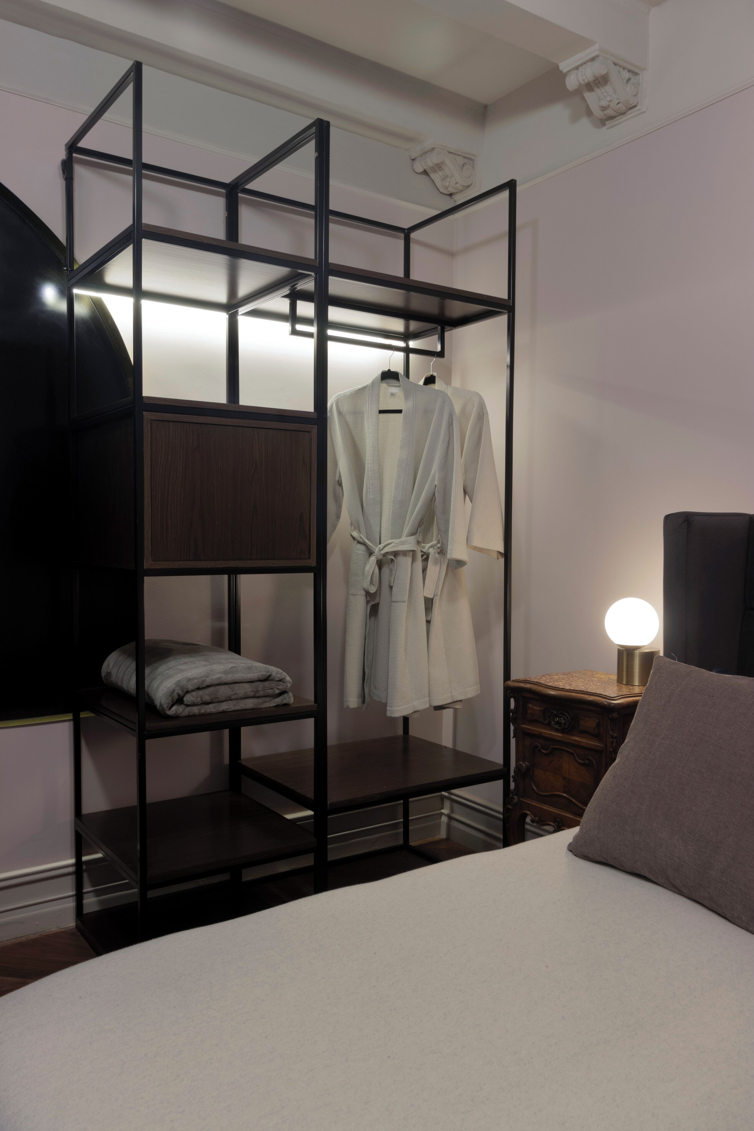 A bedroom with a bed, a wooden wardrobe, a robe hanging on the wardrobe, and a bedside lamp.