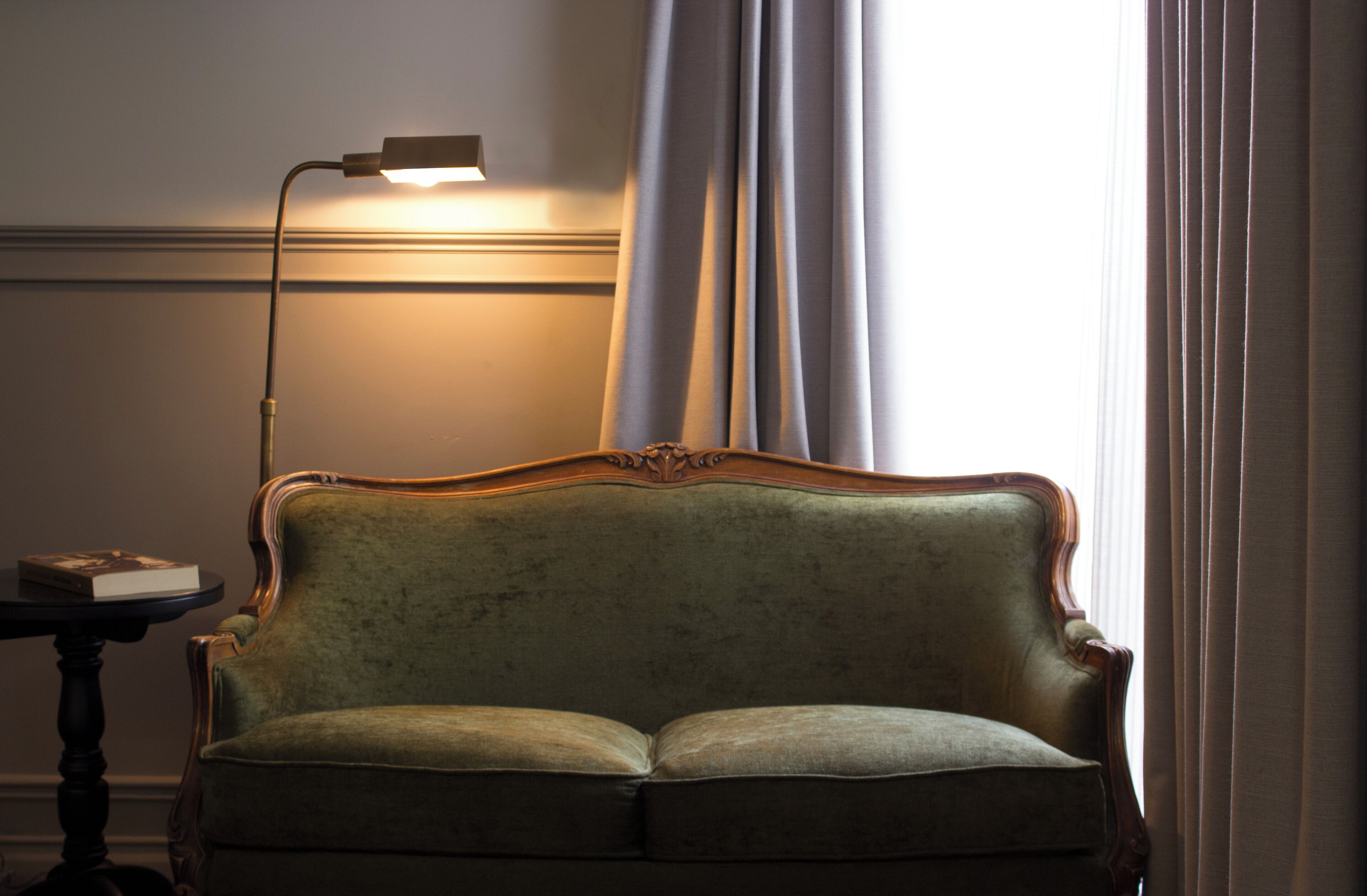 A green upholstered sofa with a wooden frame, positioned against a wall with a window and a lamp.