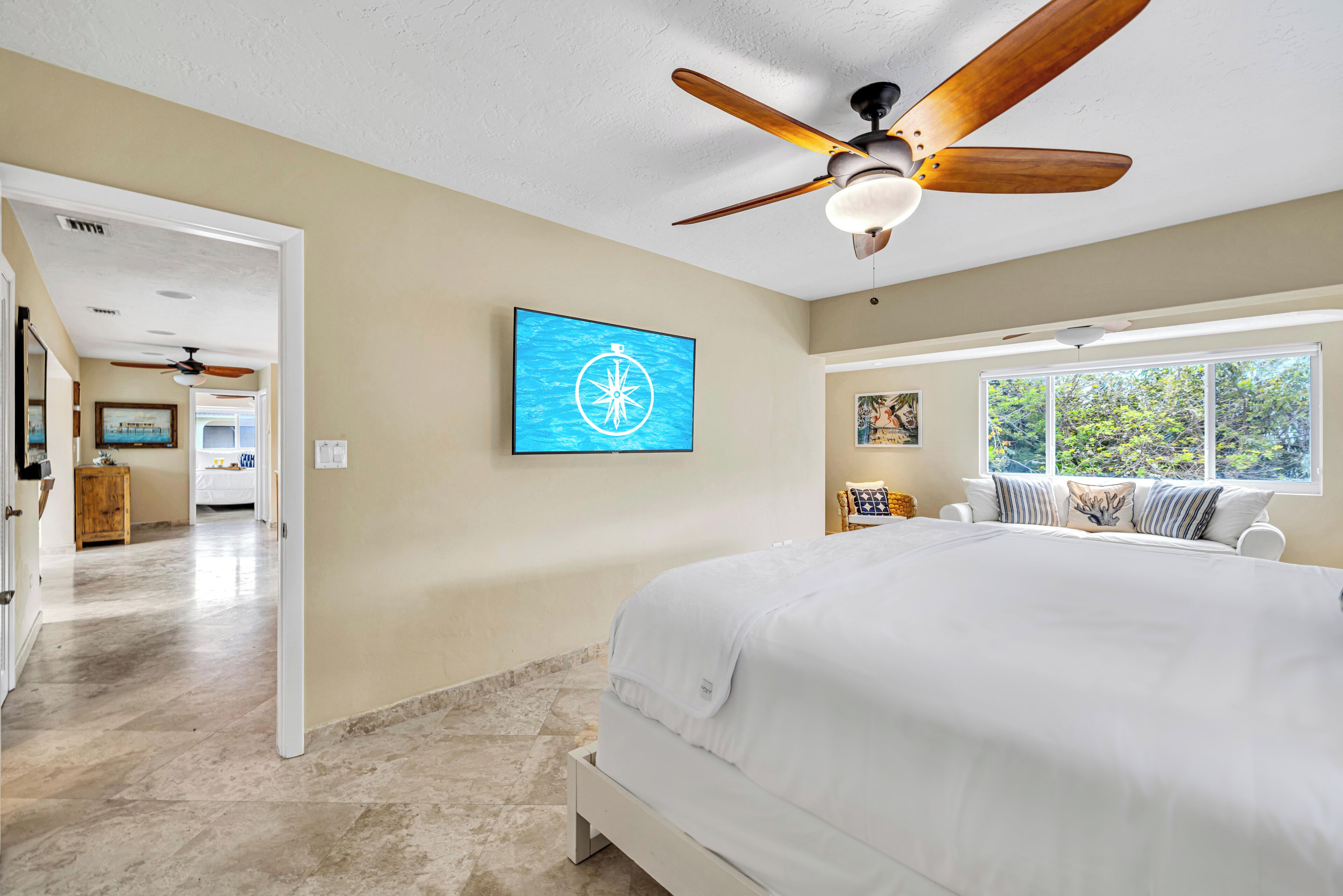 A bedroom with a large bed, a ceiling fan, a wall-mounted TV, and a window with a view of greenery.
