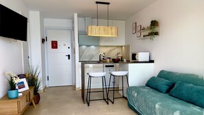 Une cuisine moderne dotée d’un four à micro-ondes encastré, d’un luminaire suspendu et d’un plan de travail blanc. On y trouve un canapé turquoise avec un coussin, une table d’appoint en bois avec un vase et un cadre photo, ainsi qu’une porte avec une enseigne rouge.