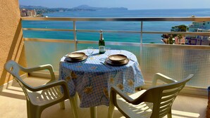 Un balcon avec une table dressée pour deux personnes, offrant une vue sur une mer turquoise et des collines lointaines.