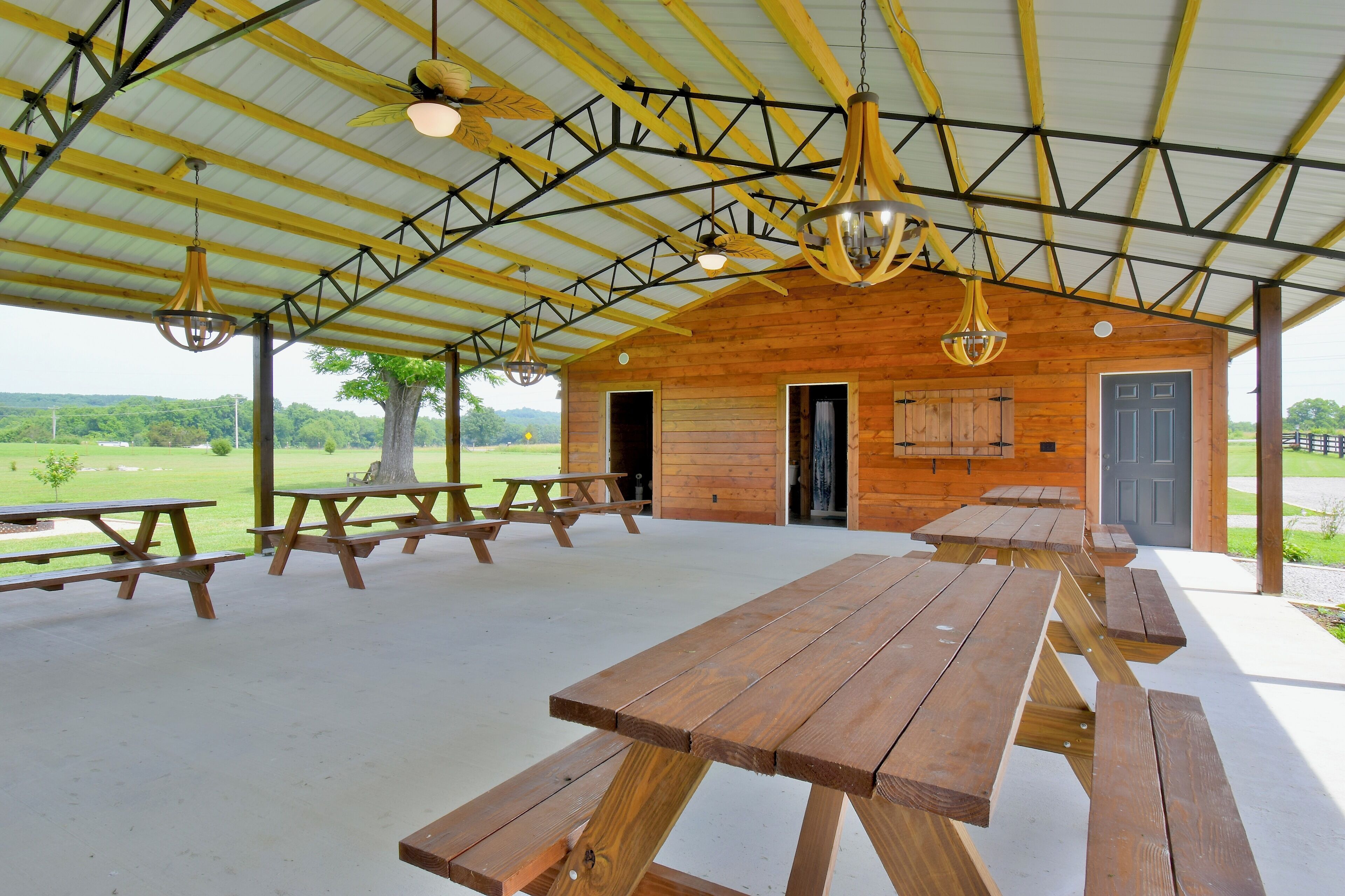 A covered picnic area with wooden benches and tables, a wooden building with a door and windows, and a view of the outdoors.