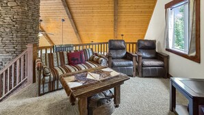 Een knus loft-achtig vertrek met een stenen muur, houten balken in het plafond, een bank, twee leren fauteuils, een salontafel en een groot raam.