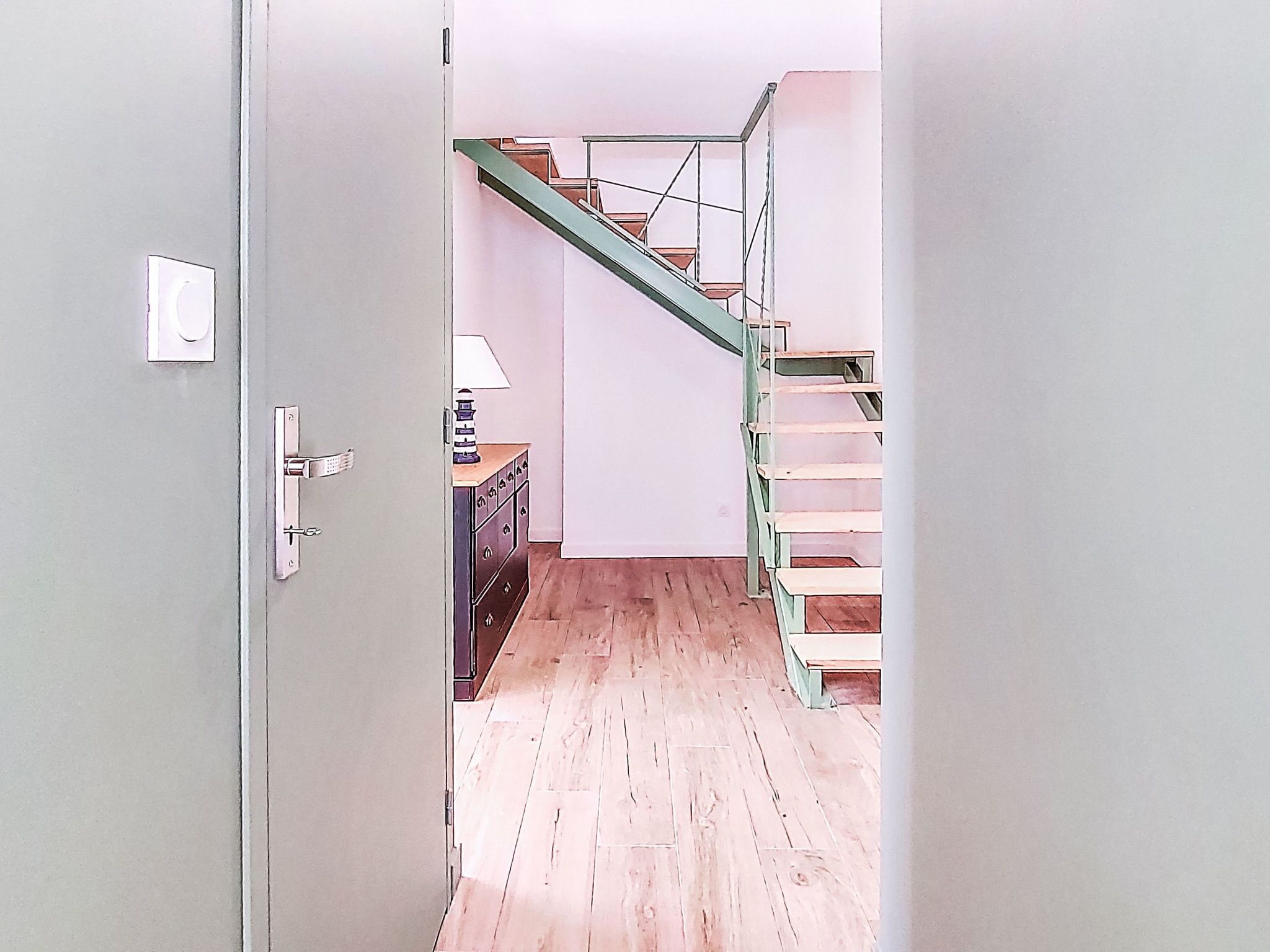A hallway with a wooden floor, a staircase with green railings, and a small table with a lamp.