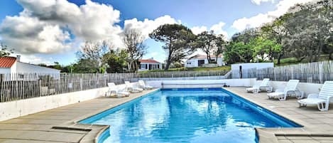 A rectangular swimming pool with a tiled deck, surrounded by white lounge chairs and a wooden fence.