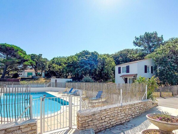A swimming pool with a wooden fence, a stone wall, and a white building with a red roof in the background.