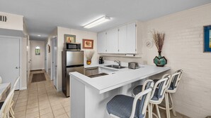 A modern kitchen with white cabinets, a stainless steel refrigerator, and a bar area with bar stools. - 2-Bedroom Beachfront Comfort w/ Heated Pool! (South Padre Island)