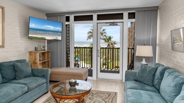A living room with a large TV, two sofas, a coffee table, and a balcony with a view of the beach and palm trees. - 2-Bedroom Beachfront Comfort w/ Heated Pool! (South Padre Island)