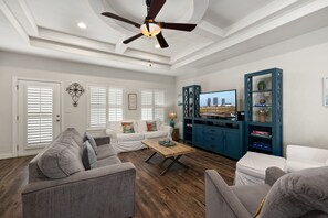 A spacious living room with a ceiling fan, white walls, and a large TV. - Beautiful Coastal Sol Villa • Heated Pool • 3 Min Walk to Beach (South Padre Island)