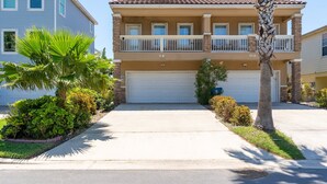 A two-story residential building with a white garage door, a balcony on the second floor, and a well-maintained front yard with palm trees and shrubs. - Beautiful Coastal Sol Villa • Heated Pool • 3 Min Walk to Beach (South Padre Island)
