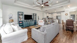A modern living room with a grey sofa, white armchair, and a TV on a stand. There is a dining area with a table and chairs, and a kitchen in the background. - Beautiful Coastal Sol Villa • Heated Pool • 3 Min Walk to Beach (South Padre Island)